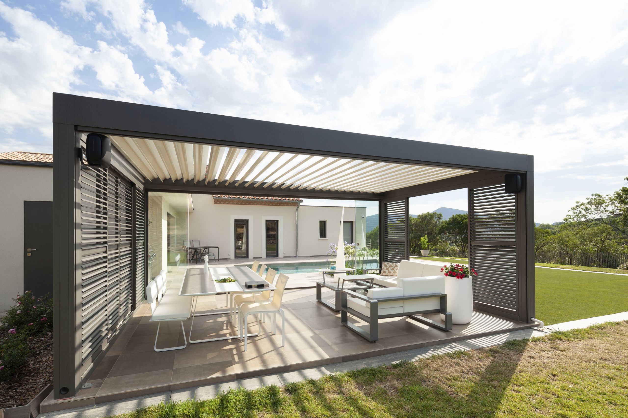 Moderne freistehende Terrasse mit Lamellendach und Ausblick auf einen einladenden Pool, umgeben von stilvollen Möbeln und grünem Rasen