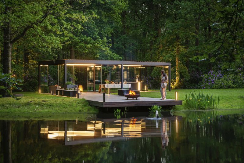 Freistehende, verglaste Terrasse, mitten im Wald. Die Terrasse hat eine indirekte Beleuchtung und wird von zwei Personen am Lagerfeuer genutzt